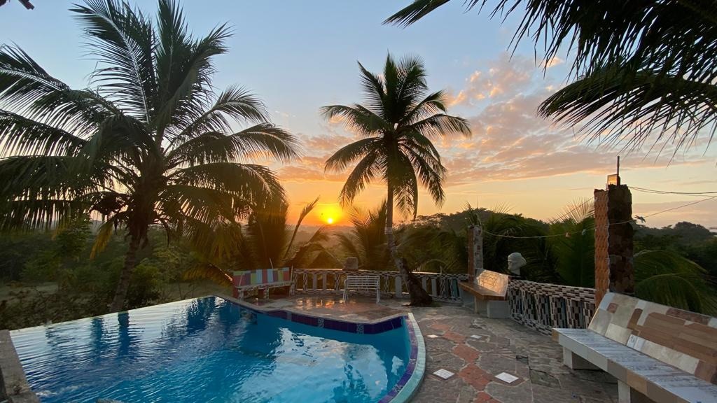 A serene pool area is framed by tall palm trees, with a vibrant sunrise visible over the horizon. The water reflects warm tones, creating a tranquil setting for relaxation. Comfortable seating options are arranged around the pool, inviting moments of peace in this private oasis.