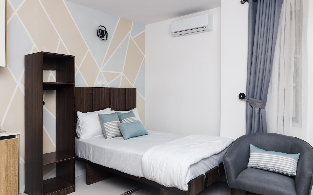A room featuring a spacious bed layered with white linens and decorative pillows in soft blue tones. Light wood furniture includes a shelf unit and a cozy armchair. A large window allows natural light to fill the space, while air conditioning is mounted on the wall.