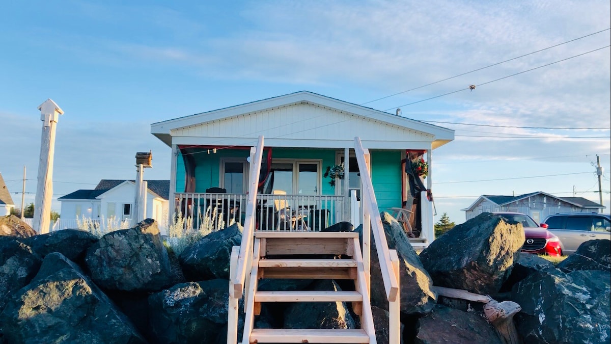 A seaside chalet is displayed, featuring a welcoming entrance with wooden steps leading to a porch. The exterior showcases a light blue color, complemented by decorative elements. Large windows provide natural light, while rocky landscaping frames the property, enhancing its coastal charm.