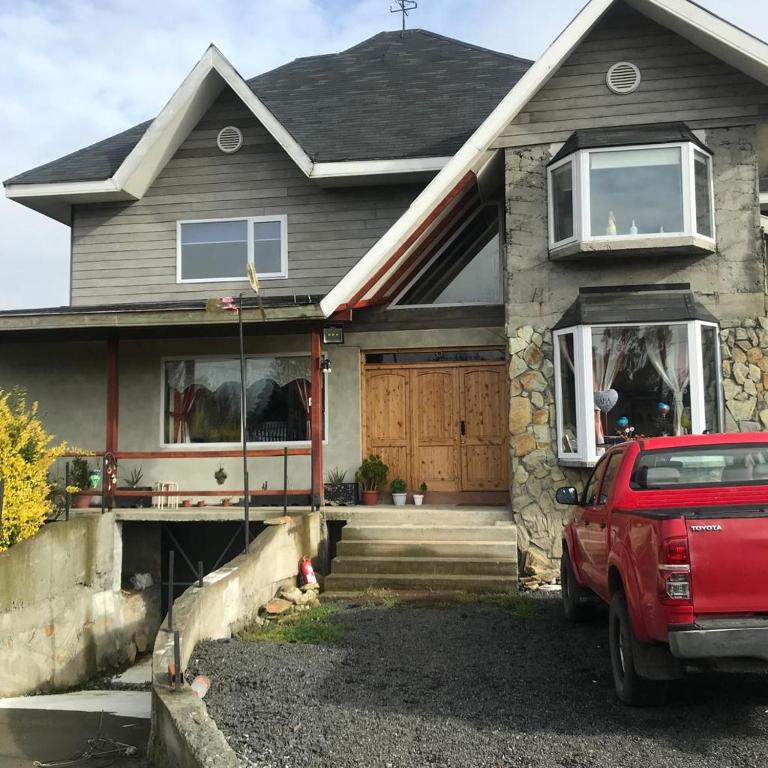 The exterior of a three-story house is displayed, featuring a mix of stone and wooden accents. Large bay windows are visible on the right, and a wooden front door is centrally positioned. A red vehicle is parked in front on a gravel surface, complemented by surrounding greenery.
