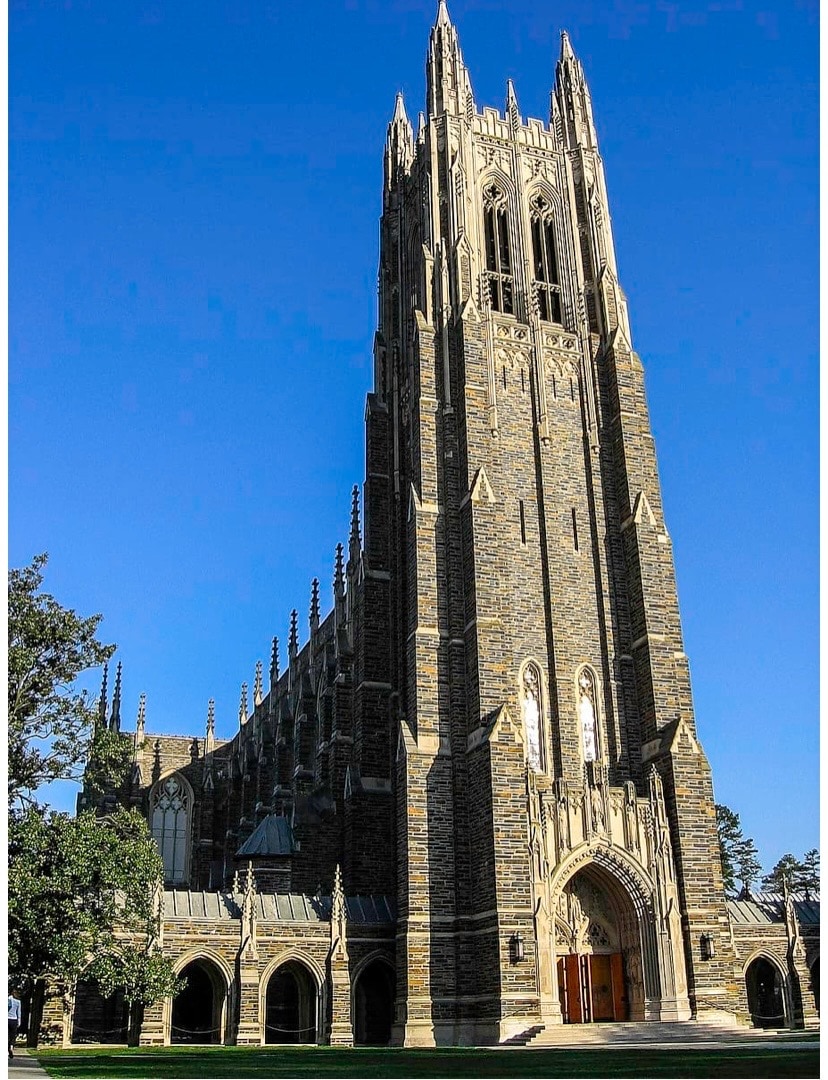 A tall stone tower rises against a clear blue sky, featuring intricate gothic architectural details and pointed spires. The entrance is marked by large wooden doors framed by arched stonework. Lush greenery surrounds the base of the building, enhancing its grandeur.