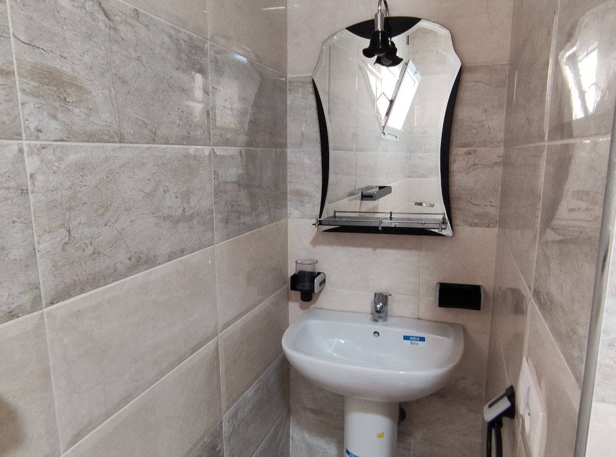 A modern bathroom is featured, with smooth grey tiles covering the walls. A compact white sink is positioned below a uniquely shaped mirror, and a soap dispenser is mounted nearby. Natural light filters through a small window, enhancing the bright and clean ambiance.
