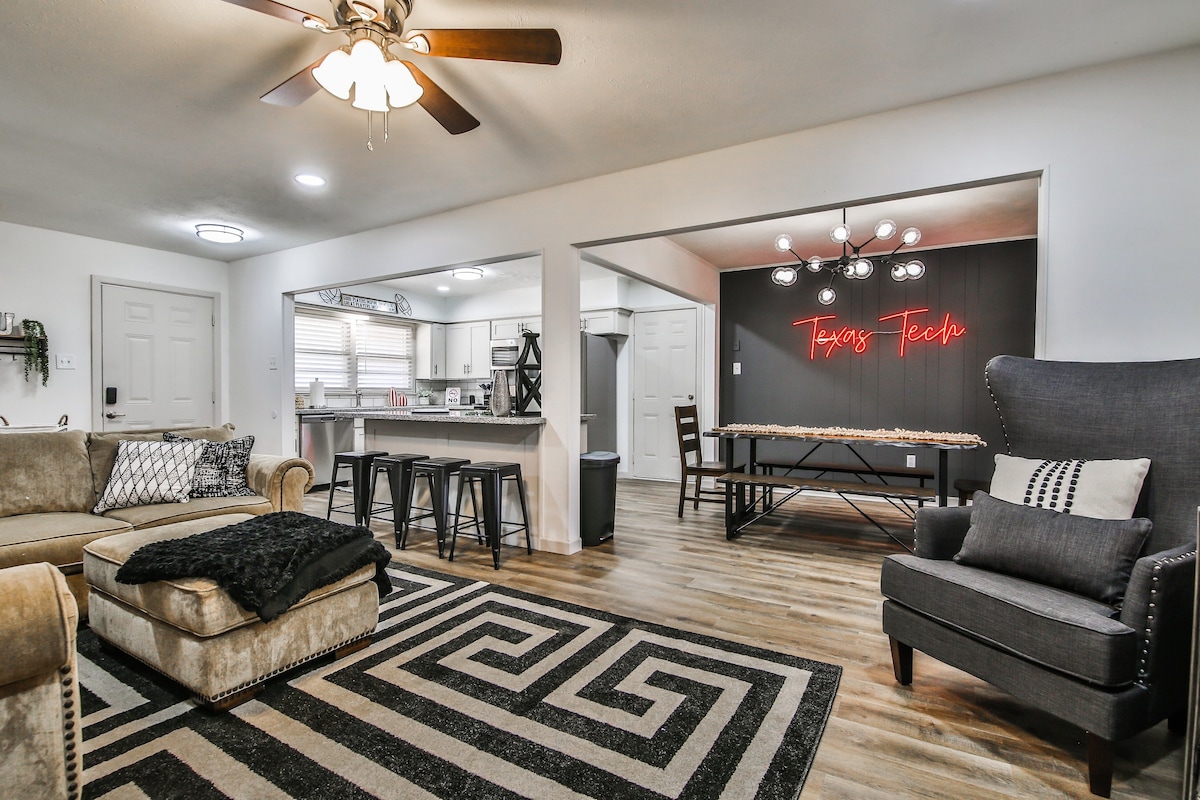 A spacious living area showcases a comfortable sofa layered with a dark throw blanket. A patterned rug defines the space, leading to a dining area with a long table and stylish chairs. The adjoining kitchen features modern appliances, complemented by a welcoming light fixture.
