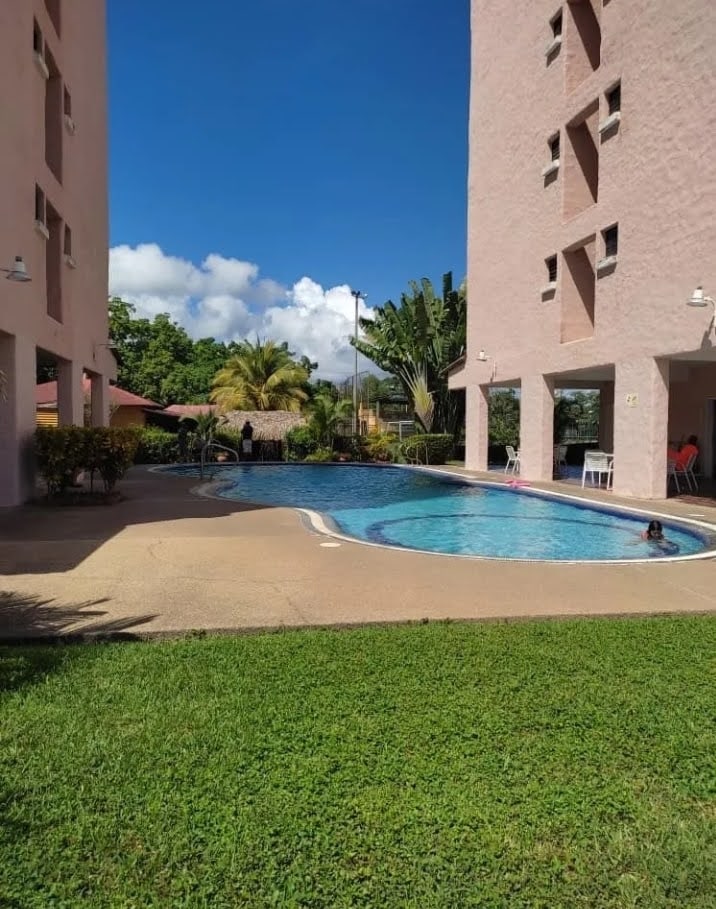 A pool area is framed by two light-colored buildings, with lush greenery and palm trees surrounding the space. Clear blue water reflects the sky, while sun loungers are positioned on the pool deck. A few guests enjoy leisure activities in and around the pool.