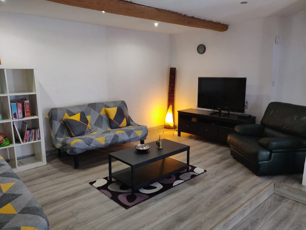 A living area features two patterned sofas with yellow accents and a coffee table at the center. A TV is positioned on a low, dark cabinet, complemented by a floor lamp providing soft illumination. A small bookshelf displays various items, enhancing the space's welcoming ambiance.