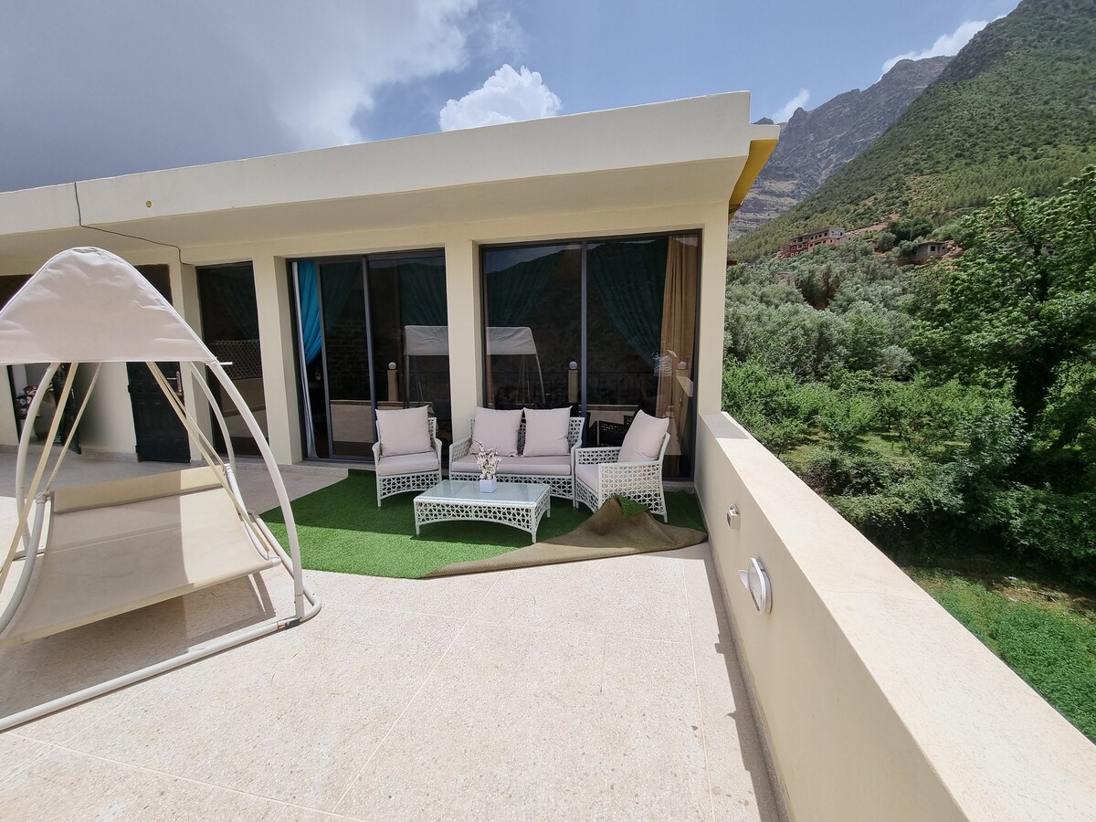 A spacious terrace is featured with comfortable seating arrangements, including a light-colored sofa and chairs. Lush greenery and mountain views are visible in the background. The area is designed for relaxation, with sliding glass doors providing access to the interior.