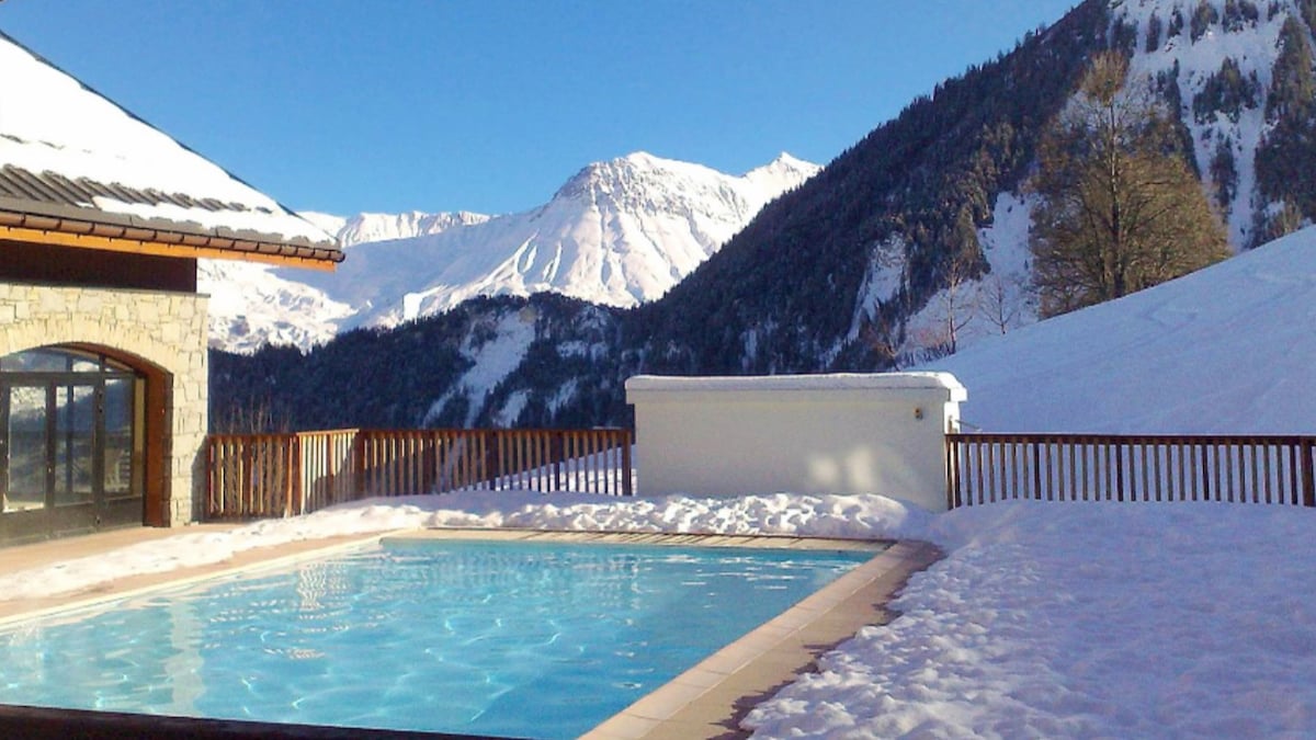 An inviting outdoor pool is surrounded by snow-covered terrain, with stunning mountain views in the background. Sunlight reflects off the pool's surface, creating a serene atmosphere. A wooden fence defines the area, enhancing the sense of seclusion.