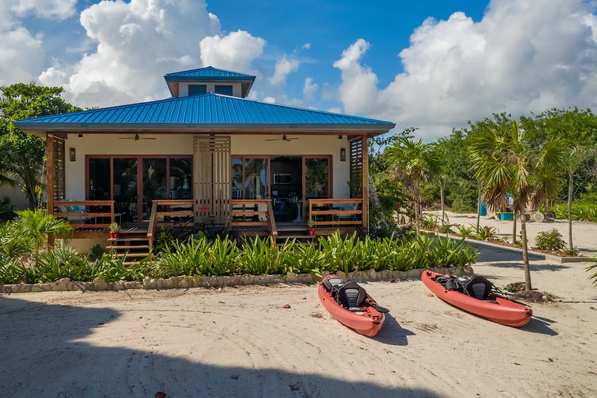 The beachfront villa is showcased with a vibrant blue roof, surrounded by lush greenery and sandy pathways. Two kayaks are positioned on the beach in front of the villa, inviting guests to enjoy water activities. Large windows suggest spacious indoor areas with plenty of natural light.