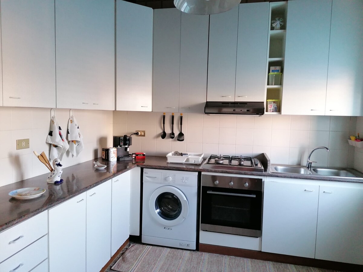 The kitchen is equipped with modern appliances, including a gas stove, oven, and washing machine. Ample counter space is available, complemented by white cabinetry. Cooking utensils hang on the wall, and a sink is positioned next to the stove, providing a functional cooking environment.