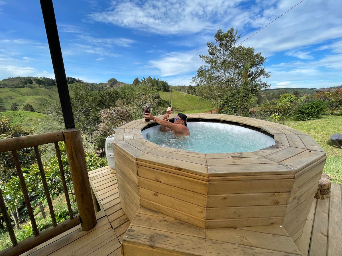 An outdoor wooden hot tub is set on a deck overlooking rolling hills and greenery. The structure is circular with a smooth, polished surface. The clear blue sky complements the serene landscape, enhancing a tranquil environment for relaxation.