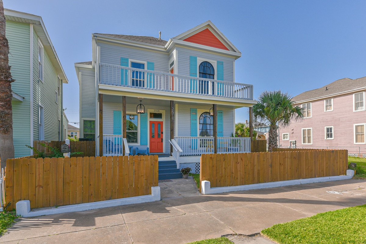 A two-story historic home is showcased with a colorful facade featuring light blue and orange accents. A spacious front porch is visible, complemented by white railings. The property is enclosed by a low wooden fence, with palm trees and neighboring houses visible in the background.