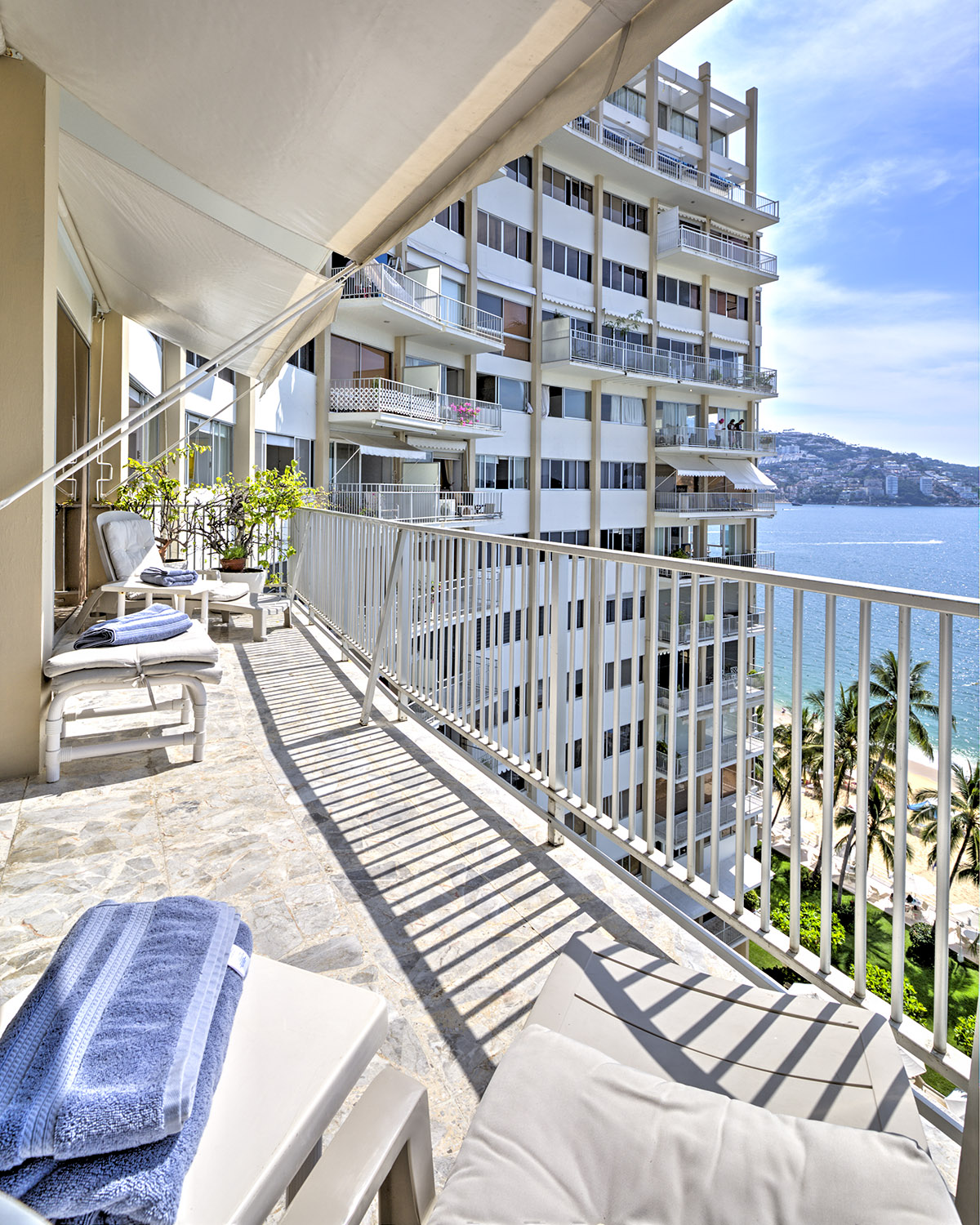 A spacious balcony is presented, featuring light-colored loungers and rolled towels. Sunlight casts shadows on the stone floor, while the surrounding building and palm trees can be seen. The bay view beyond adds a sense of openness, contributing to a relaxing atmosphere.