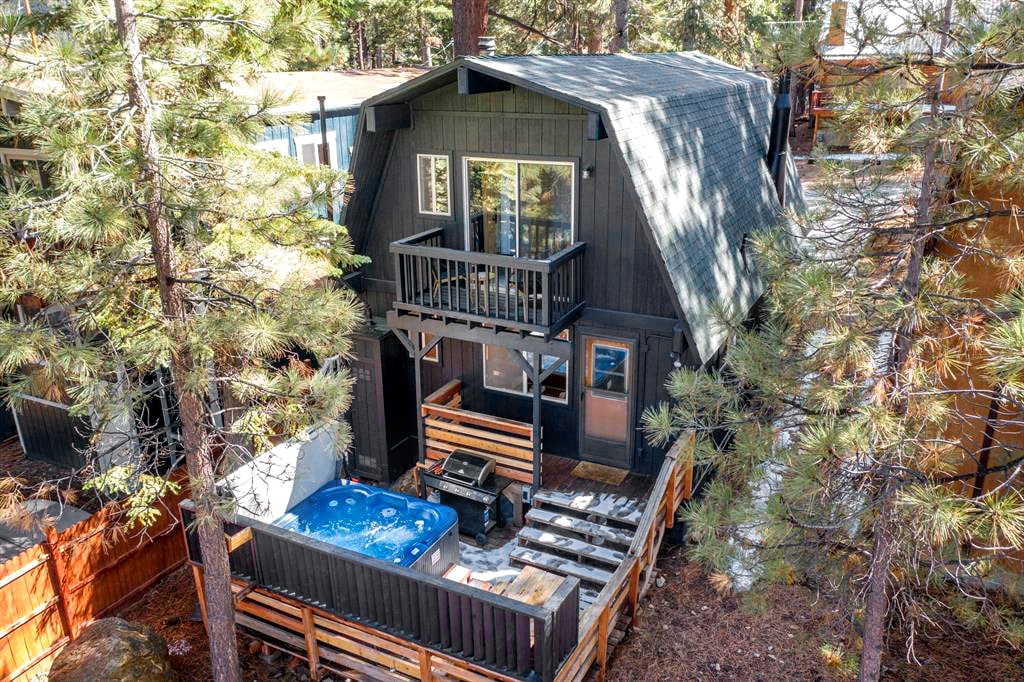 The cabin's exterior showcases a classic Gambrel A-frame design, surrounded by tall pine trees. A private balcony with sliding glass doors complements the inviting hot tub area on the deck, providing a tranquil setting for relaxation.