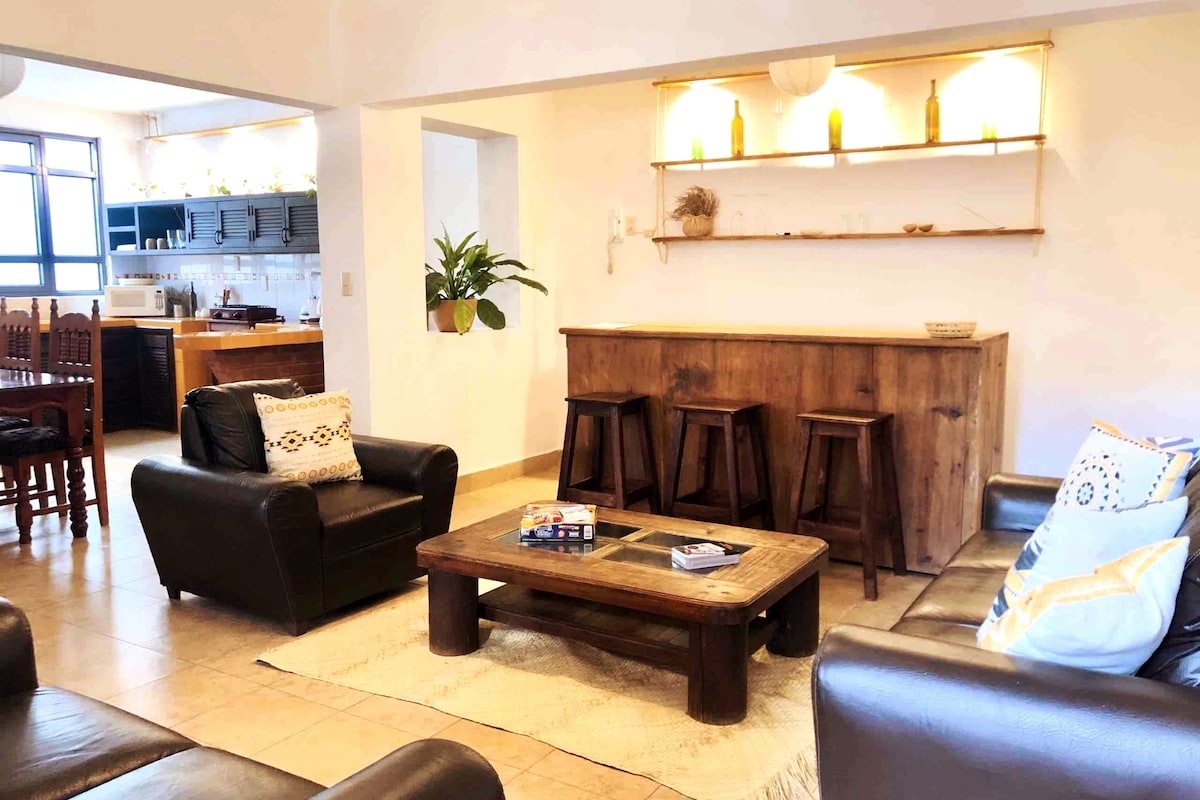 A spacious living area features dark leather sofas arranged around a wooden coffee table. A natural fiber rug sits beneath the furniture. Behind the seating, a wooden bar with three stools is highlighted, along with soft lighting and decorative items placed on shelving.