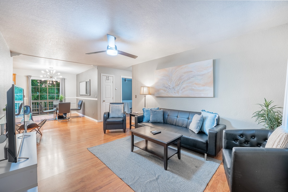 A spacious living area features hardwood floors, a dark leather sofa, and a sleek coffee table. Natural light streams in from nearby windows, while a ceiling fan adds comfort. A dining space with a table and chairs is visible in the background, enhancing the room's inviting feel.