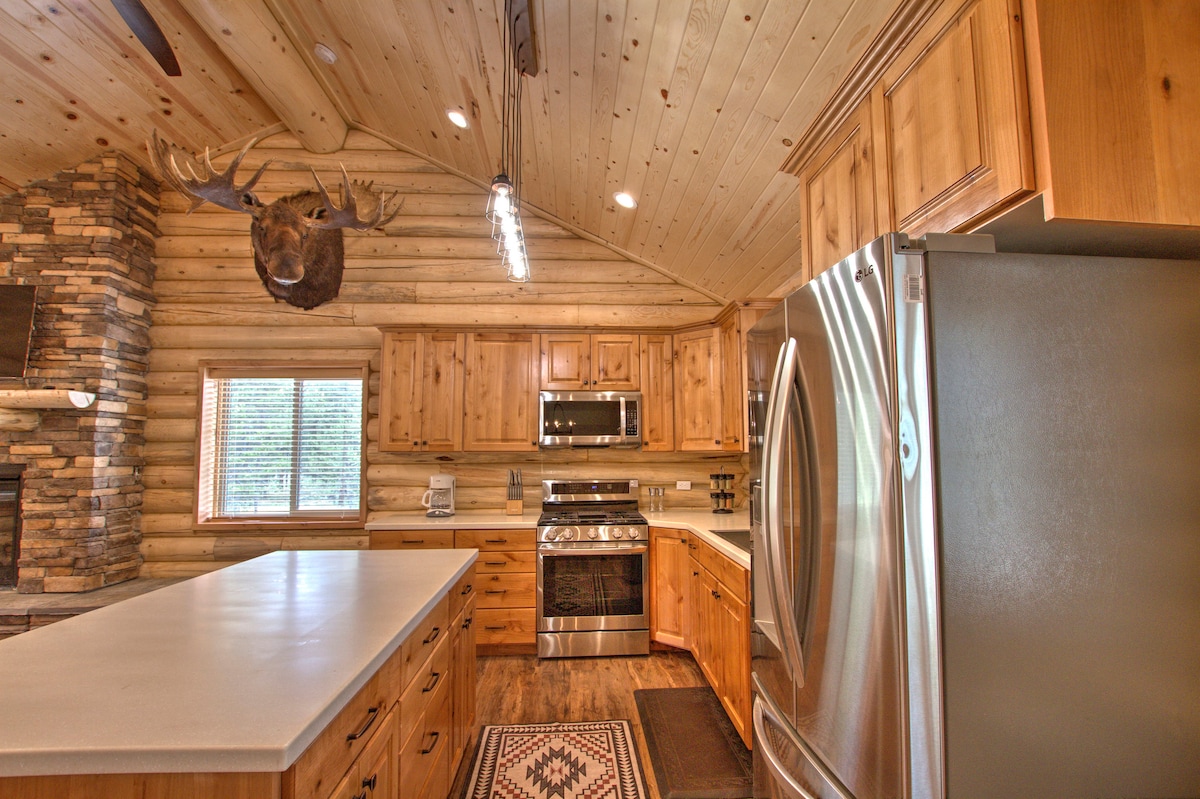 A spacious kitchen features wooden cabinetry and a large island with a light countertop. Modern stainless-steel appliances, including a refrigerator and gas stove, are present. Natural light enters through the window, emphasizing the warm wood paneling and rustic decorative elements.