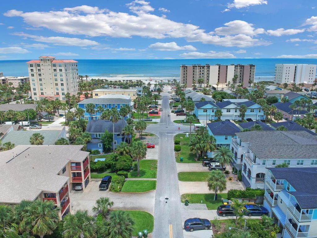 An aerial view captures the vibrant coastal community of Jacksonville Beach, featuring palm-lined streets that lead towards the ocean. Various buildings, including residences and commercial properties, are visible, with the beach and ocean stretching out in the distance under a partly cloudy sky.