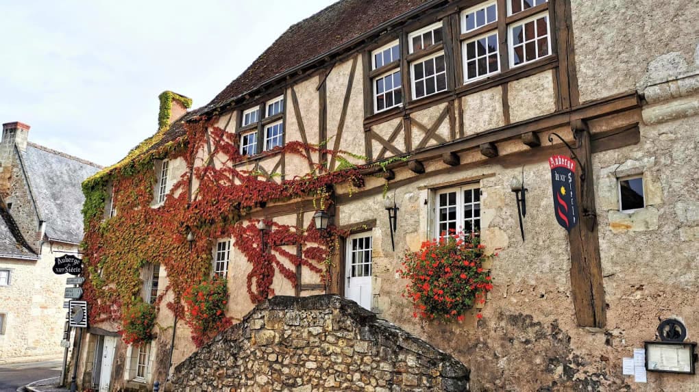 An historic building is seen featuring traditional architecture with wooden beams and stone walls. Colorful vines climb the exterior, adding a touch of nature. Flower boxes are positioned beneath the large, multi-pane windows, enhancing the charming appearance of the home.