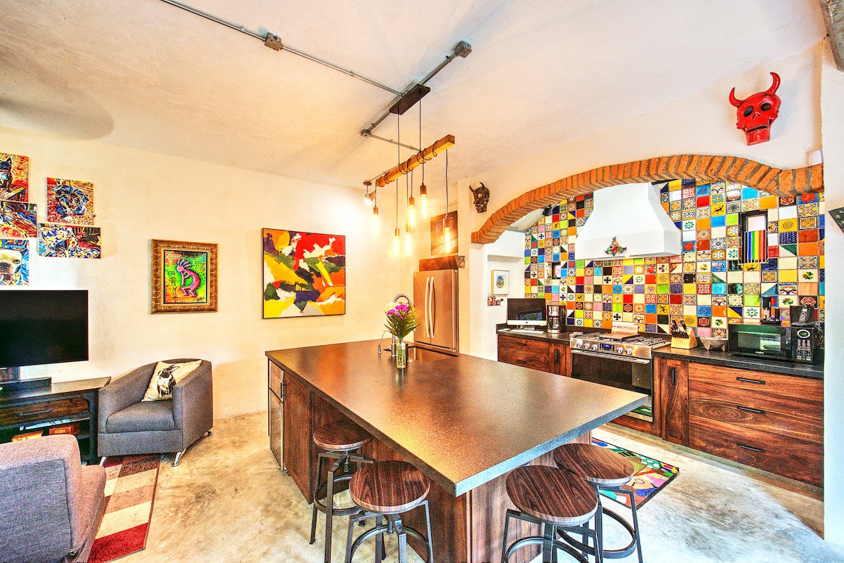 A modern kitchen features a large island surrounded by bar stools. Colorful tiles create a vibrant backsplash behind the stove. Artworks adorn the walls, and a television is positioned near a comfortable seating area on the side.