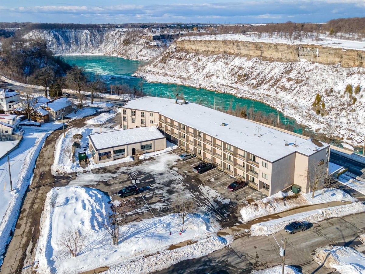 An aerial view captures the River Rapids Inn, situated near the stunning Niagara River and gorge. The snow-covered landscape surrounds the hotel, which features a spacious parking lot. The natural turquoise waters of the river stand in contrast to the winter scenery.