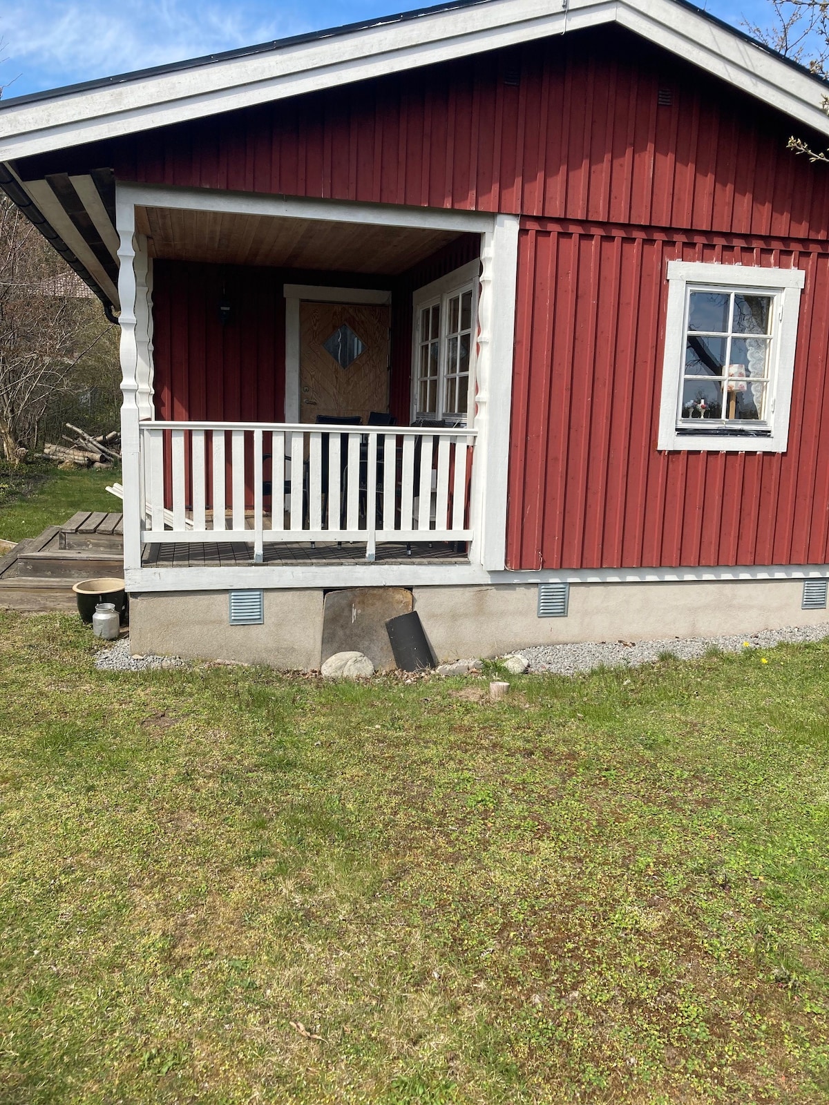 A charming red cottage features a covered front porch with white railings. Large windows allow natural light to enter. The lawn is green and well-kept, with a slight slope leading to a path beside the house. A peaceful setting is suggested by the surrounding trees.