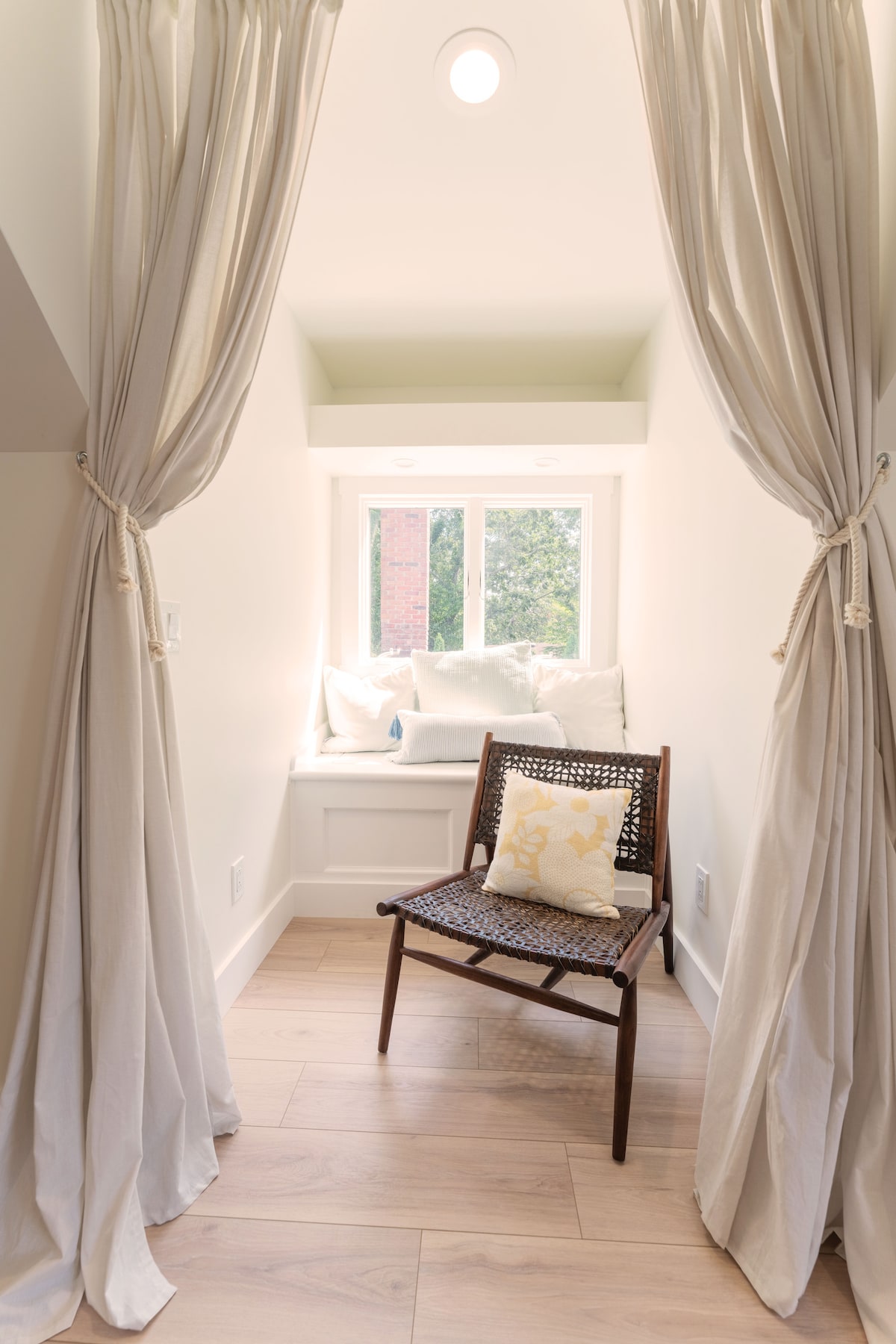 A cozy nook is created with soft, billowy curtains framing a window seat. A single chair with a woven seat is positioned nearby, while the gentle light filters in, illuminating the serene atmosphere. Plush cushions invite relaxation in this tranquil space.