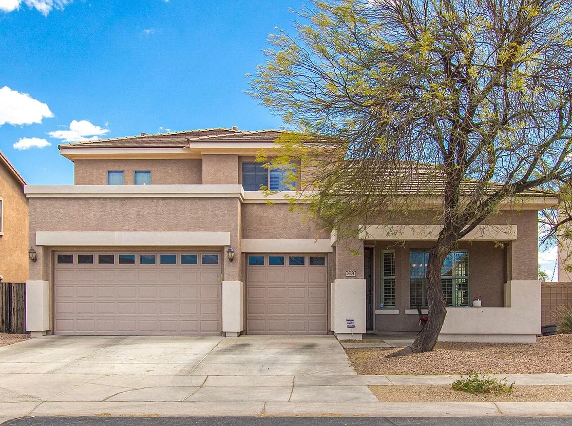 The exterior of a two-story home is visible, featuring a beige façade and large windows. A front porch is situated to the right, adorned with potted plants. Two garages are present, showcasing space for vehicles. A tree offers shade in the foreground.