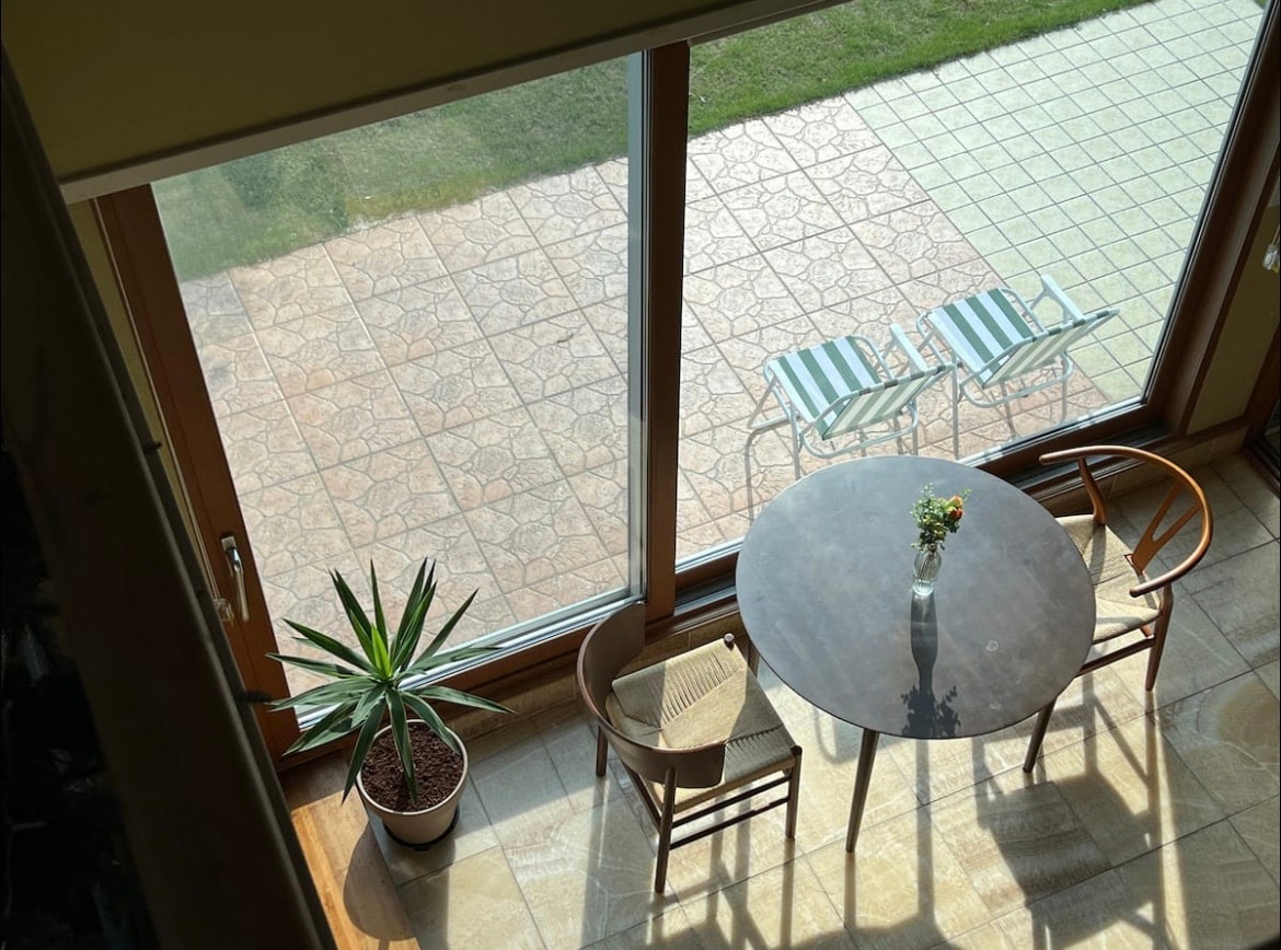 A small dining area is positioned near a large glass window, revealing a view of a grassy outdoor space. A round table with a vase of flowers is complemented by two wooden chairs. Sunlight streams in, enhancing the inviting ambiance.