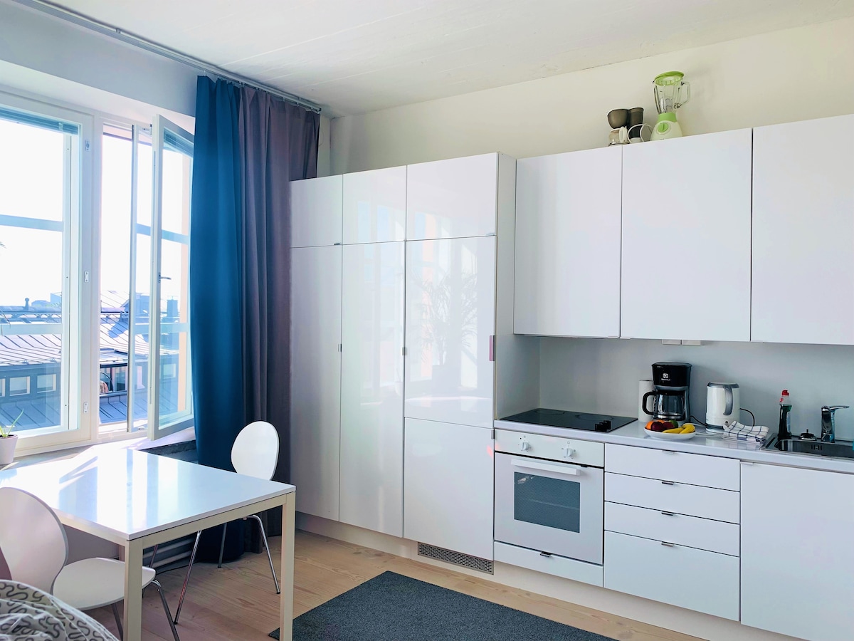 A modern kitchen area is visible, featuring sleek white cabinetry and a glass-top stove. A compact dining table with white chairs accommodates two. Natural light enters through large windows, creating an airy ambiance. A small mat adds warmth to the light wooden floor.