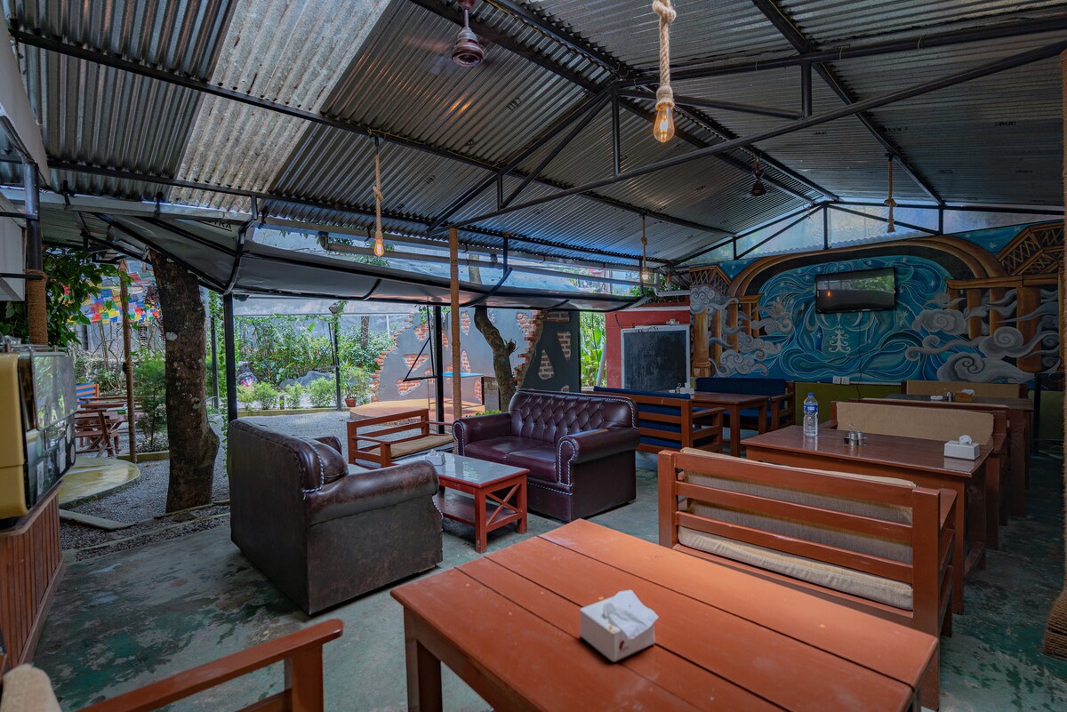 An open communal space features rustic wooden tables and a mix of seating options, including leather sofas. Natural light filters in through an overhead structure, illuminating the surrounding greenery, with vibrant wall art visible in the background.