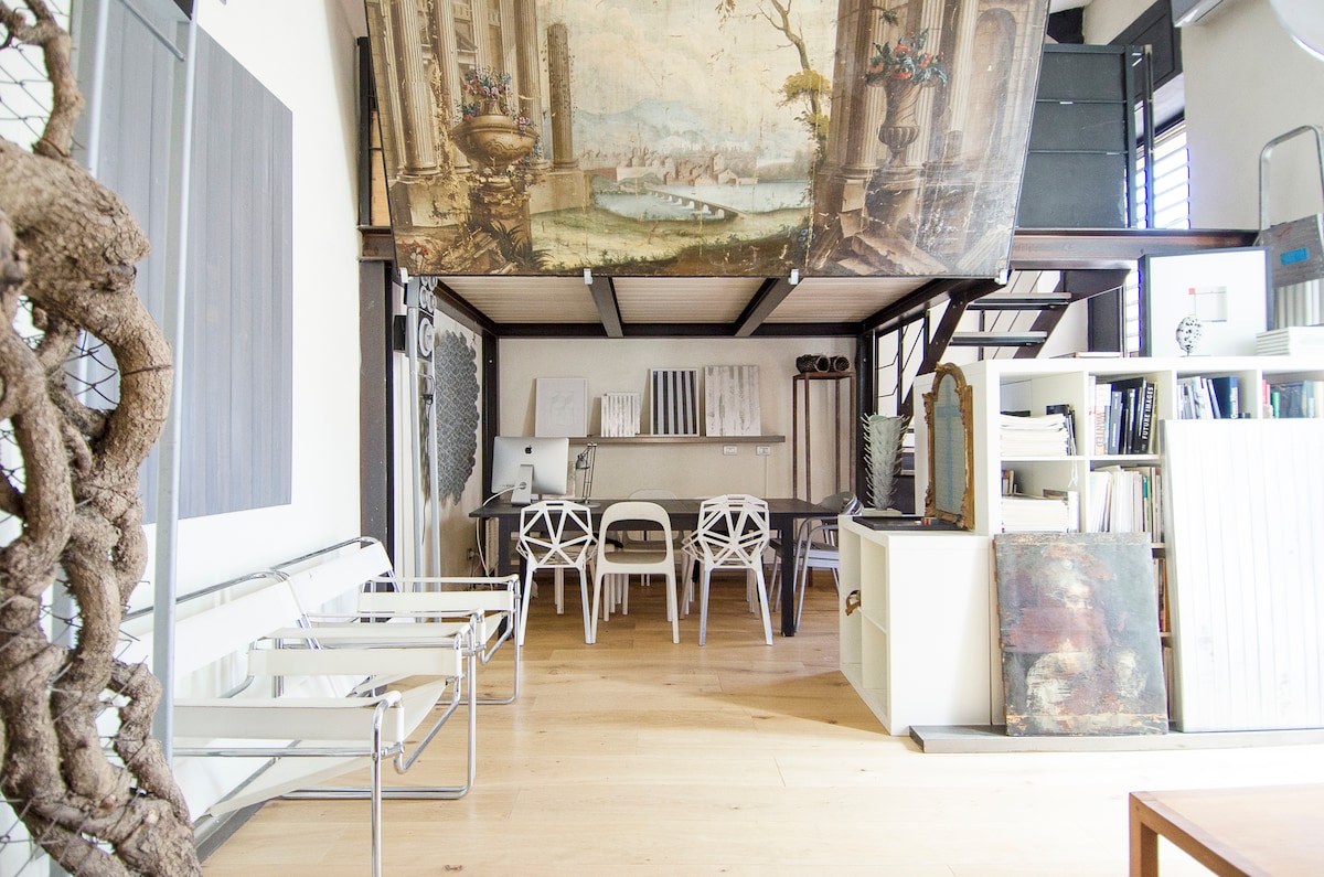 An open living space features a wooden beam ceiling and an oversized artwork suspended above. A dining area is provided with a sleek table surrounded by white chairs. Natural light illuminates the wooden floor, enhancing the contemporary design elements throughout the loft.