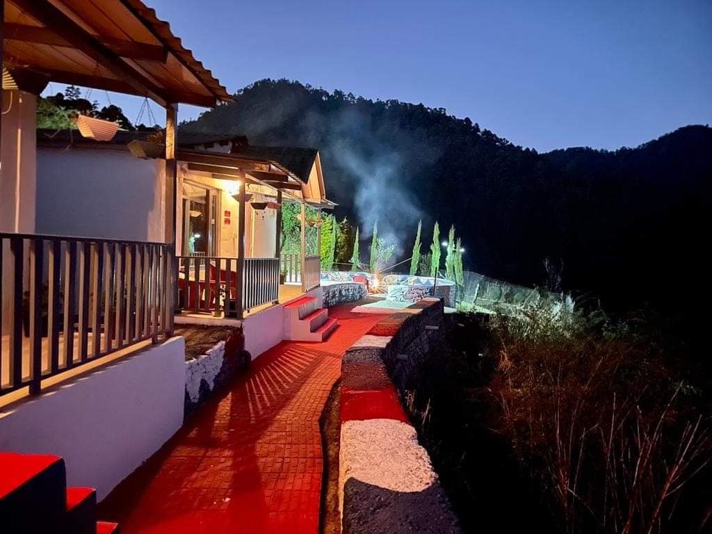 A stone pathway leads through a gently lit outdoor area, bordered by lush greenery. The warm glow from a cottage illuminates the surrounding landscape at dusk, with distant mountains creating a serene backdrop under a twilight sky.