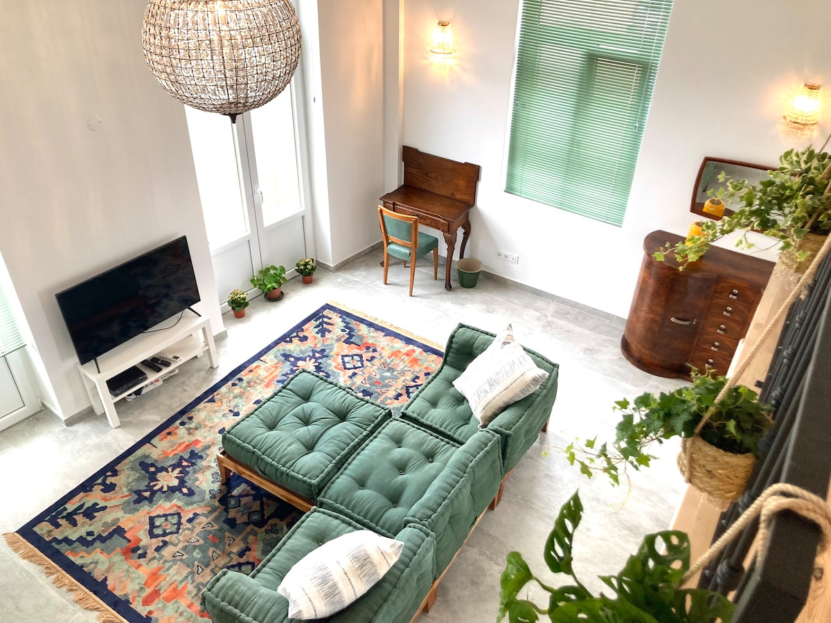 A spacious living area features a green sectional sofa with comfortable cushions, a large patterned rug, and a wooden writing desk. Natural light enters through mirrored windows, while potted plants add a touch of greenery. Soft lighting from wall fixtures complements the bright atmosphere.