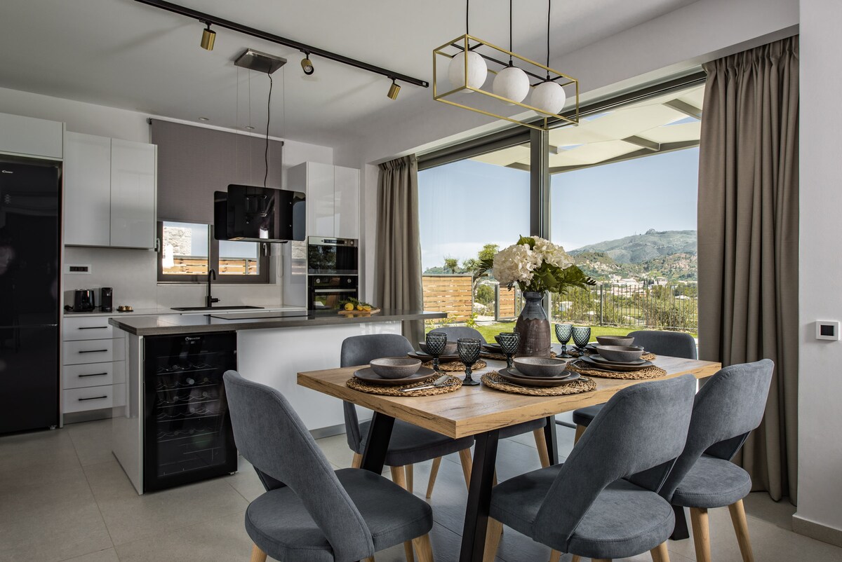 A modern dining area is featured with a wooden table surrounded by six gray chairs. Table settings include plates and a centerpiece with flowers, adding a touch of elegance. Large windows allow natural light to fill the space, offering views of the surrounding landscape.