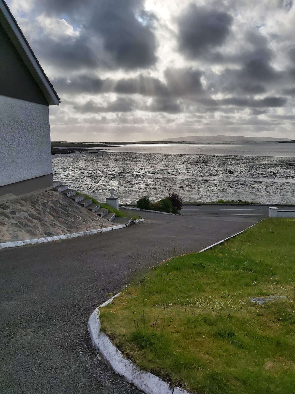 A view of the waterfront is captured from the property, showcasing a winding driveway leading towards the water. Light reflects on the waves, while clouds hover above, creating a serene atmosphere. Lush green areas border the driveway, providing a natural setting.