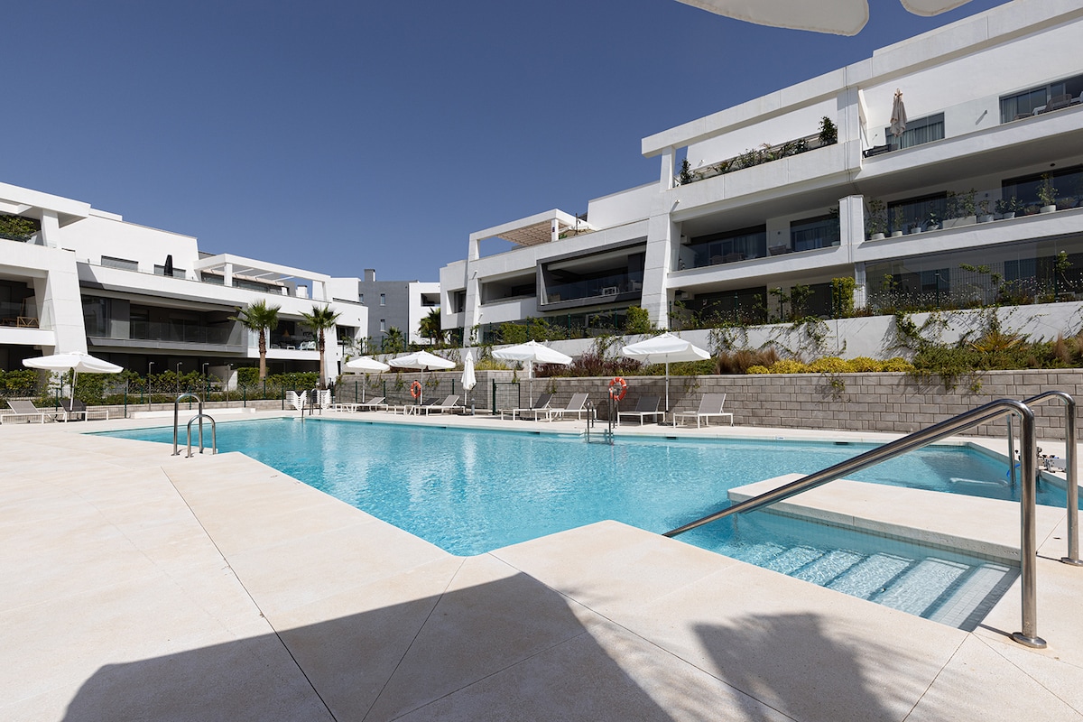 A well-maintained outdoor pool area is surrounded by modern apartment buildings. Lounge chairs and umbrellas provide shade for relaxation. The pool's turquoise waters are inviting, and steps lead down into the water, ensuring accessibility for all guests.