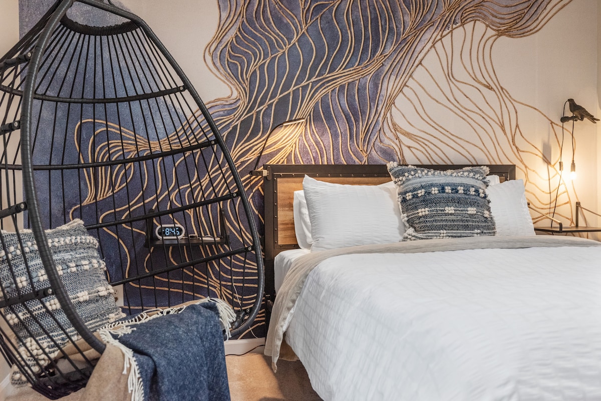 A cozy bedroom setting is highlighted by a spacious bed adorned with crisp white linens and decorative pillows. A close by hanging chair offers a unique seating option, while a softly lit lamp adds warmth to the space. A textured wall mural enhances the room's visual appeal.