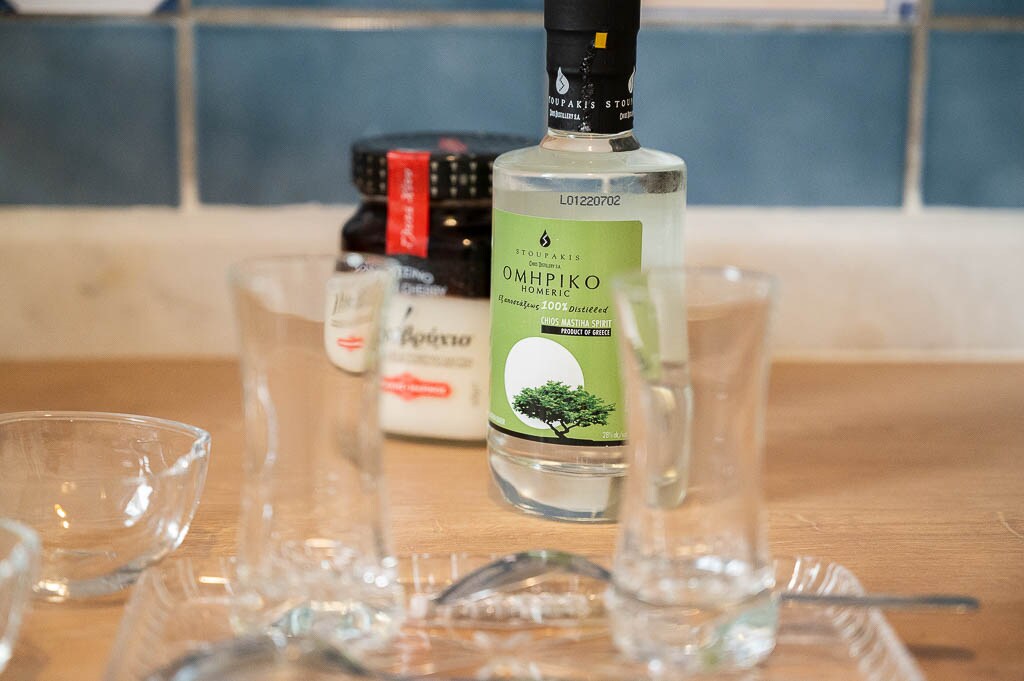 A selection of glassware is displayed on a wooden surface, accompanied by a bottle of Ouzo featuring a green label. A jar is visible behind the bottle, adding to the dining setup. The backdrop includes blue tiles, enhancing the inviting ambiance.