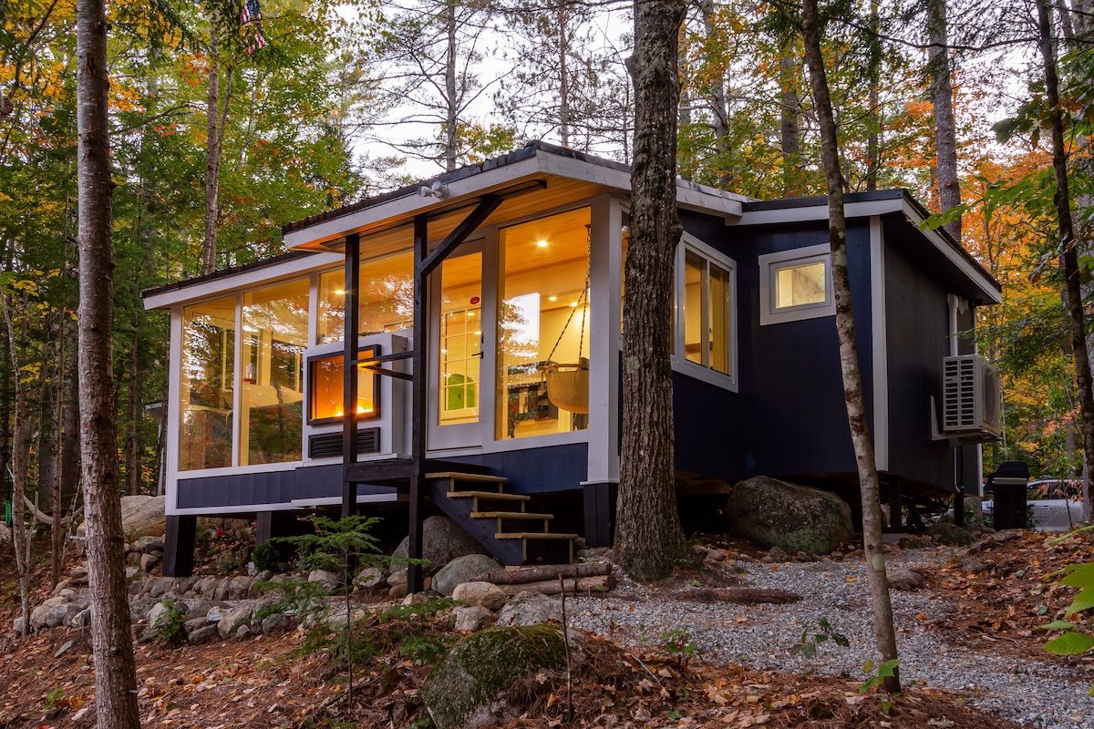 A modern cabin is nestled among trees, featuring large glass windows that invite nature inside. The structure is elevated on a stone foundation and showcases a blend of dark siding and warm wood accents. Steps lead up to an inviting entrance.