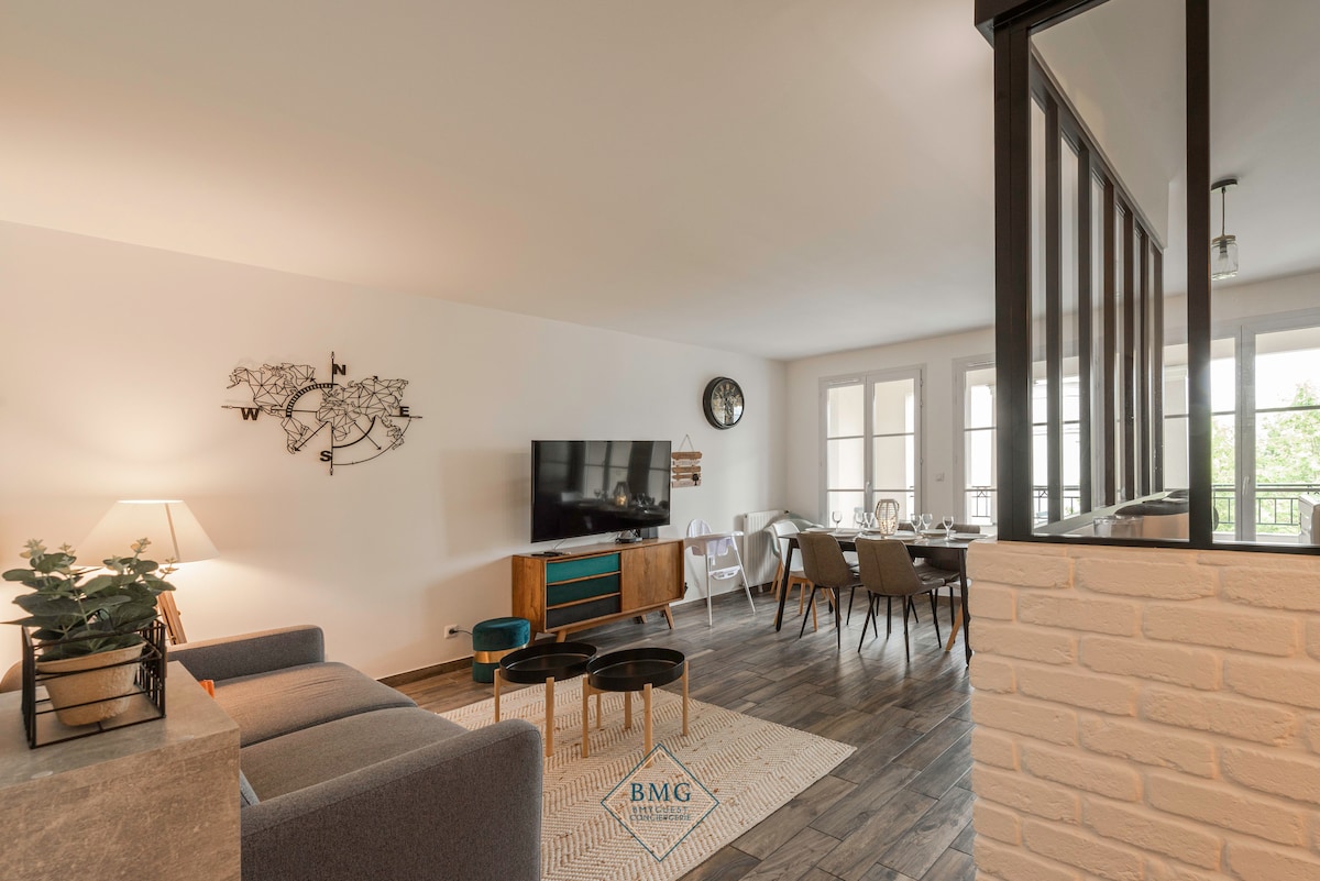 A bright living area features a comfortable sofa paired with small side tables. A television is mounted on the wall, while a dining table sits nearby. Large windows allow natural light to fill the space, and a clock adds a functional touch.