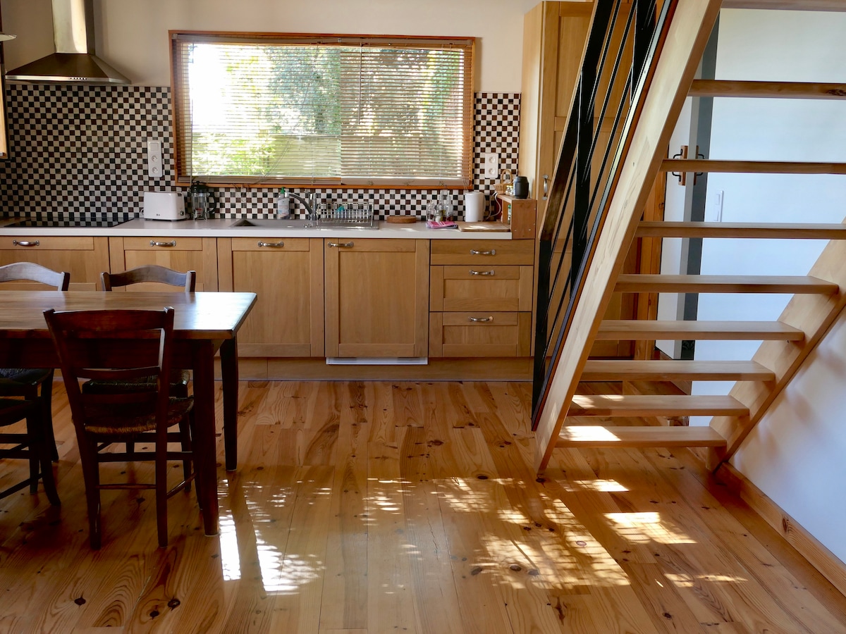 A bright kitchen features wooden cabinetry and a checkered backsplash. Natural light highlights the polished wooden floor. A dining table with matching chairs occupies part of the space, while a staircase with wooden steps blends with the overall design.
