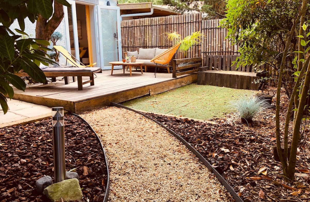 A serene garden space features a wooden deck with two lounge chairs and a small table. A winding gravel path leads through the landscaped area, showcasing vibrant greenery and minimalist decor. A small bush and ornamental grass provide subtle accents among the natural elements.