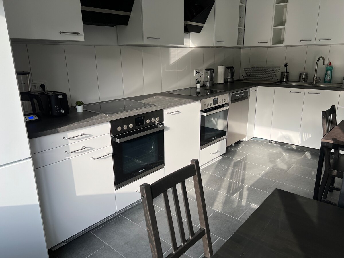 A fully equipped kitchen space features modern appliances including an oven, stovetop, microwave, and dishwasher. Dark countertops contrast with white cabinetry. A small plant adds a touch of greenery. Black wooden chairs surround a dining table, providing seating for meal preparation and enjoyment.