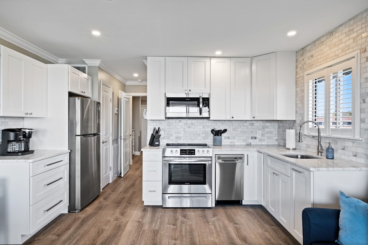 The newly renovated kitchen features sleek white cabinetry, marble countertops, and stainless steel appliances. Amenities include a full-size refrigerator, oven, and built-in microwave. Natural light filters through the window, illuminating the contemporary design and spacious layout.