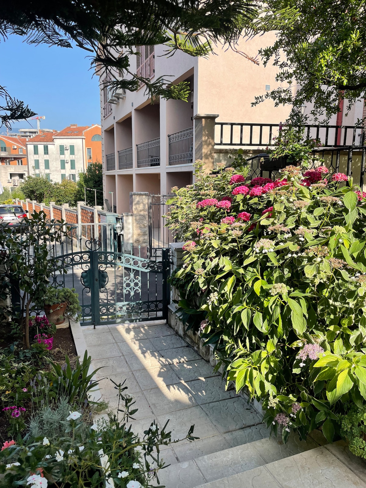 A serene pathway leads to a gated entrance, surrounded by lush greenery and vibrant flowering plants. The soft, natural light illuminates the area, enhancing the inviting atmosphere of the outdoor space. The surrounding buildings offer a calm backdrop to the garden setting.