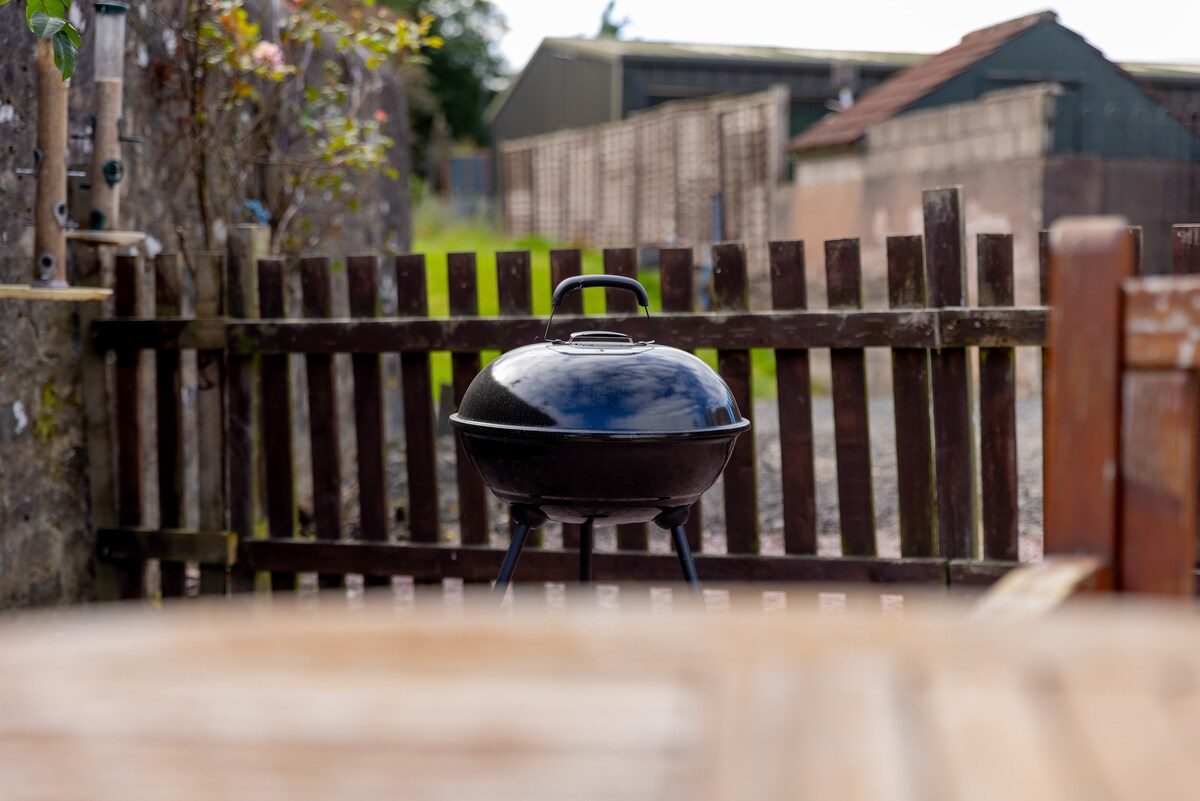 A black charcoal grill is positioned behind a wooden picket fence, surrounded by greenery. The rustic setting of a farm can be observed in the background, with buildings stretching out on either side. Natural light adds a clear view of the grilling area.