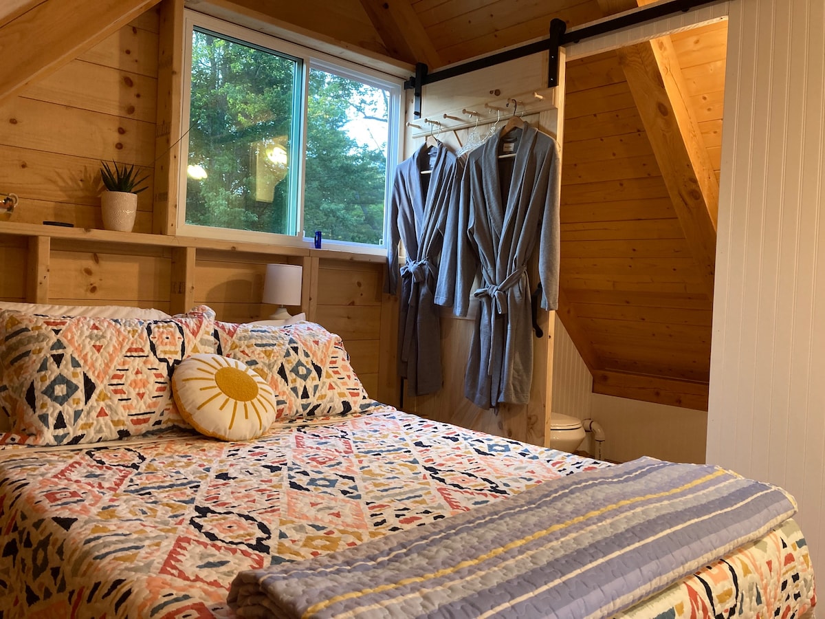 A cozy sleeping area features a colorful quilted bedspread and a decorative pillow. Cotton bathrobes hang on a rail, providing convenience. Natural light filters through the window, illuminating the wooden walls and creating a warm atmosphere.
