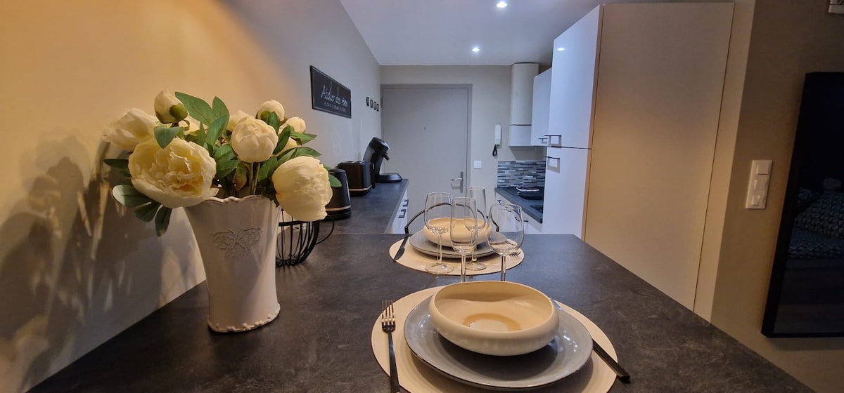 A functional dining area is presented, featuring a dark countertop with neatly arranged tableware. A decorative vase holds fresh flowers, while soft lighting enhances the space. The kitchen, visible in the background, includes modern appliances and ample storage.