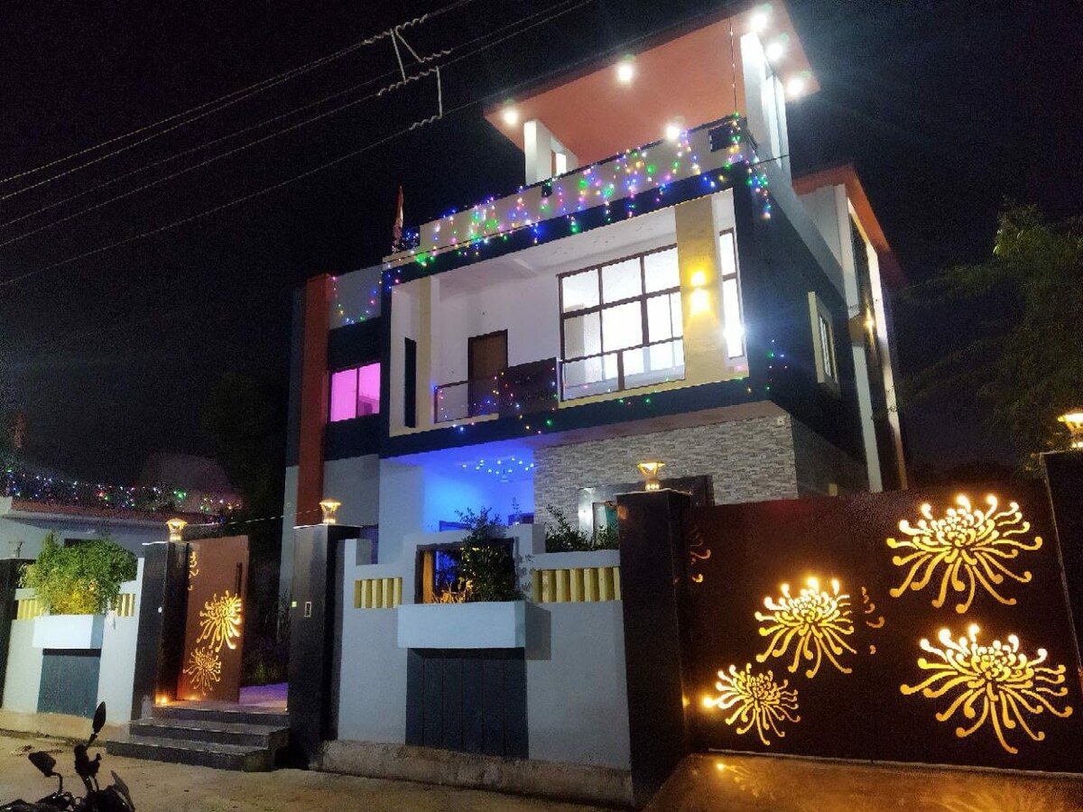 The exterior of a modern, three-story house is illuminated at night, showcasing colorful lights along the balcony. A decorative gate features intricate floral patterns, while well-maintained landscaping is visible in front. The house's windows reflect vibrant hues, enhancing its contemporary design.