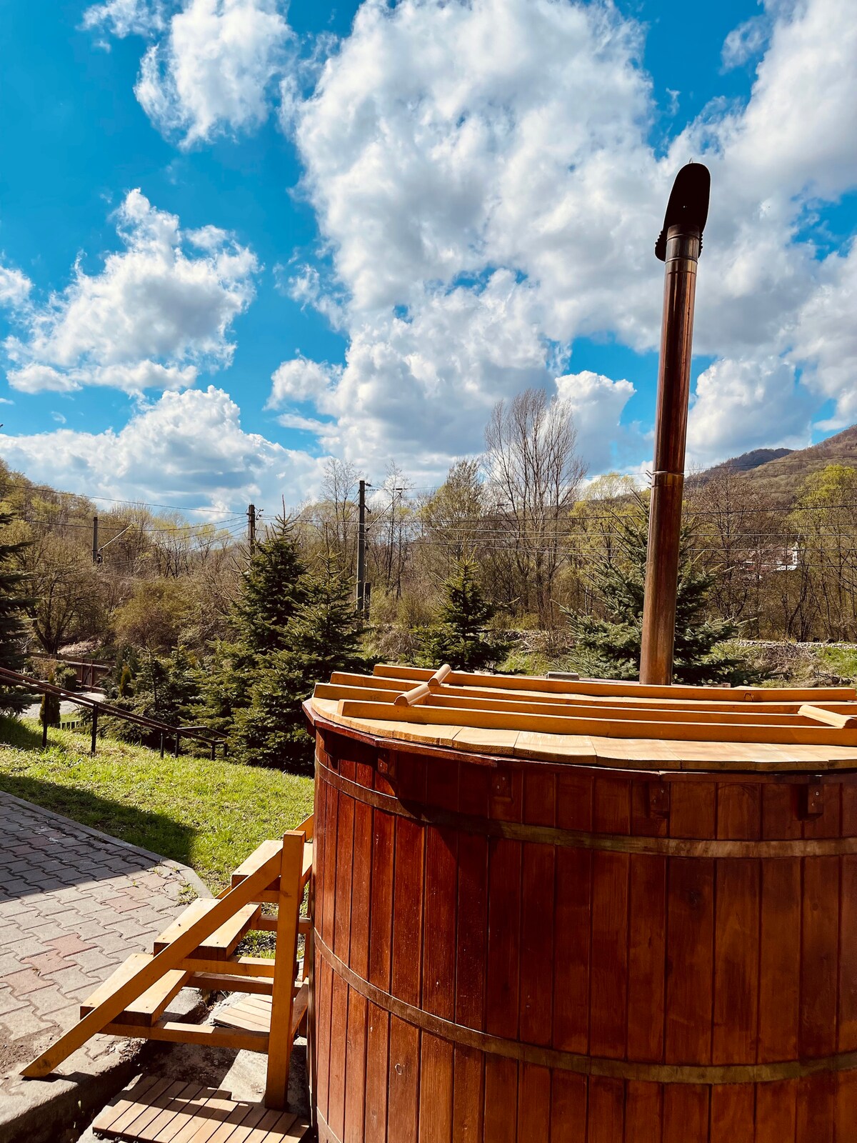 A wooden hot tub is placed outdoors, surrounded by lush greenery and trees. A chimney extends from the tub, and blue skies with scattered clouds create a serene backdrop.